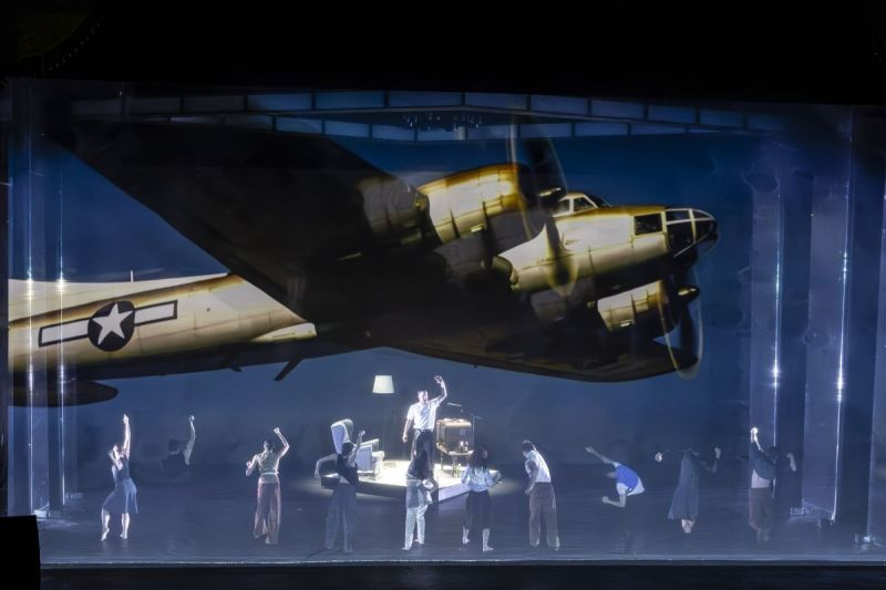 Foto di scena. Cantante in centro. In primo piano dei ballerini. Sullo sfondo viene proiettato un aereo da guerra