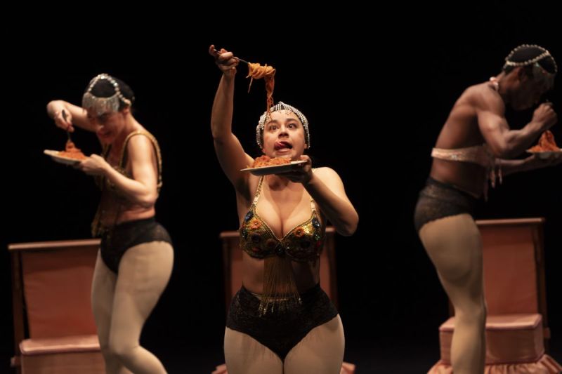 Foto di scena. Ballerine/attrici che si muovo sul palco mangiando spaghetti