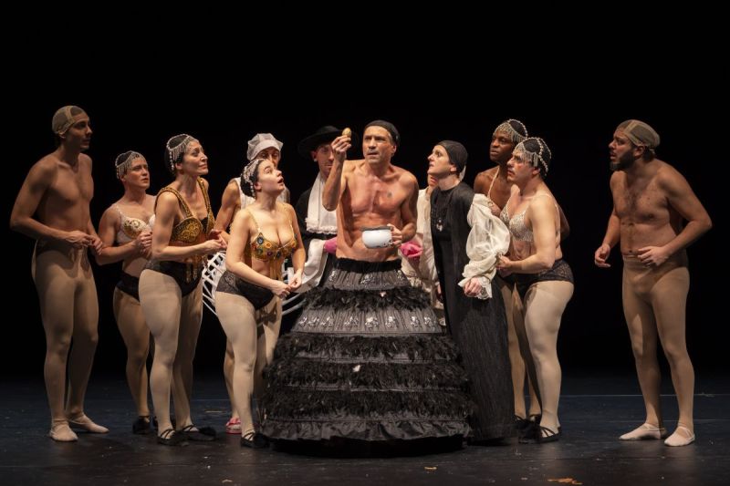 Foto di scena. Re Chicchinella guarda un oggetto. Attorno a lui tutti i suoi servitori lo osservano