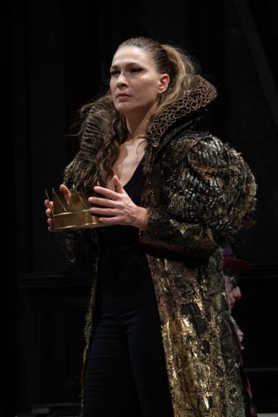 Foto di scena. Una donna in piedi tiene in mano una corona con sguardo solenne 