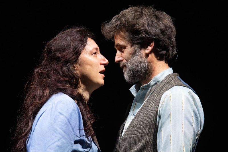 Foto di scena. Un uomo e una donna si parlano stando uno di fronte all'altra. Sfondo nero 