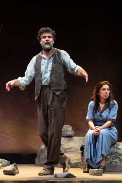 Foto di scena. In primo piano un uomo in piedi. Dietro di lui una donna vestita di blu seduta su un masso