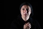 Foto di scena. Primo piano su donna vestita di nero su sfondo nero