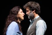 Foto di scena. Un uomo e una donna si parlano stando uno di fronte all'altra. Sfondo nero 