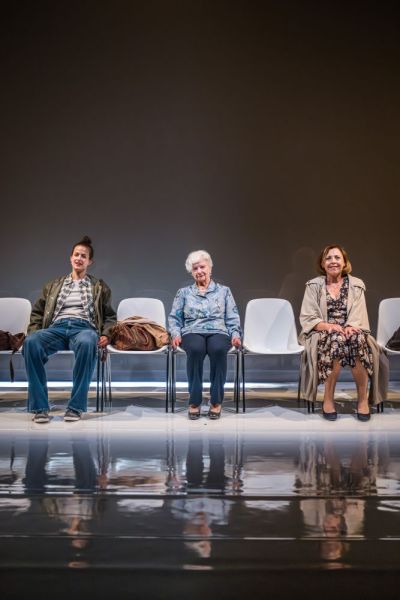 Foto di scena. Ariella Reggio, Maria Ariis e Lucia Limonta sedute su una fila di sedie guardano verso il pubblico