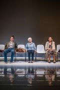 Foto di scena. Ariella Reggio, Maria Ariis e Lucia Limonta sedute su una fila di sedie guardano verso il pubblico